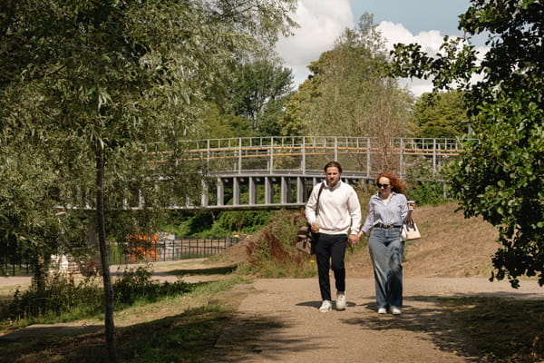 Male and female walking along dirt path near riverside with arch bridge in background