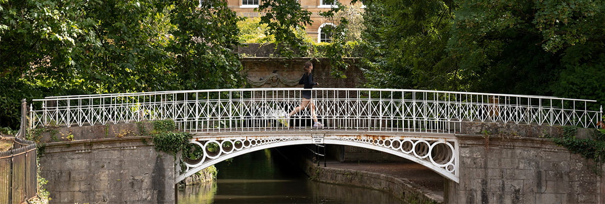 Female jogging along arch bridge over river with trees and building in background