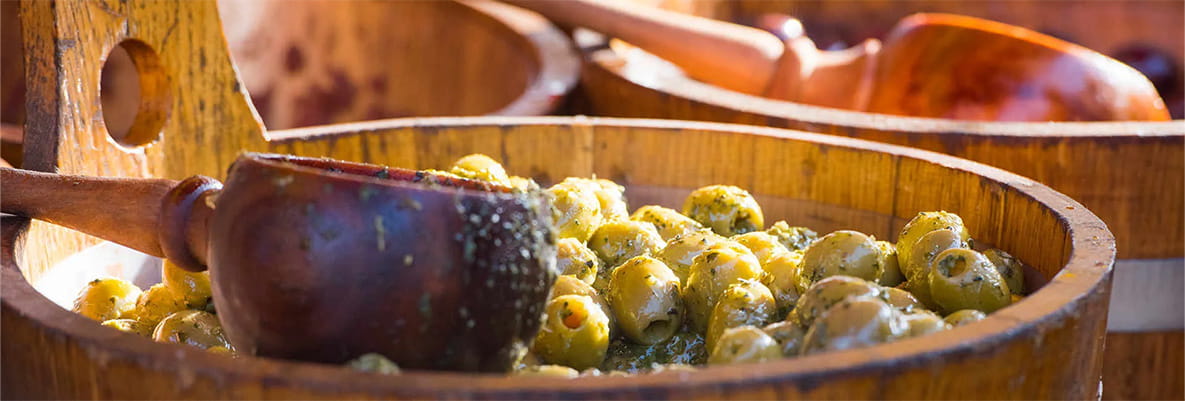 A close-up shot of green olives marinated in herbs and oil, displayed in a rustic wooden bowl with a wooden serving spoon