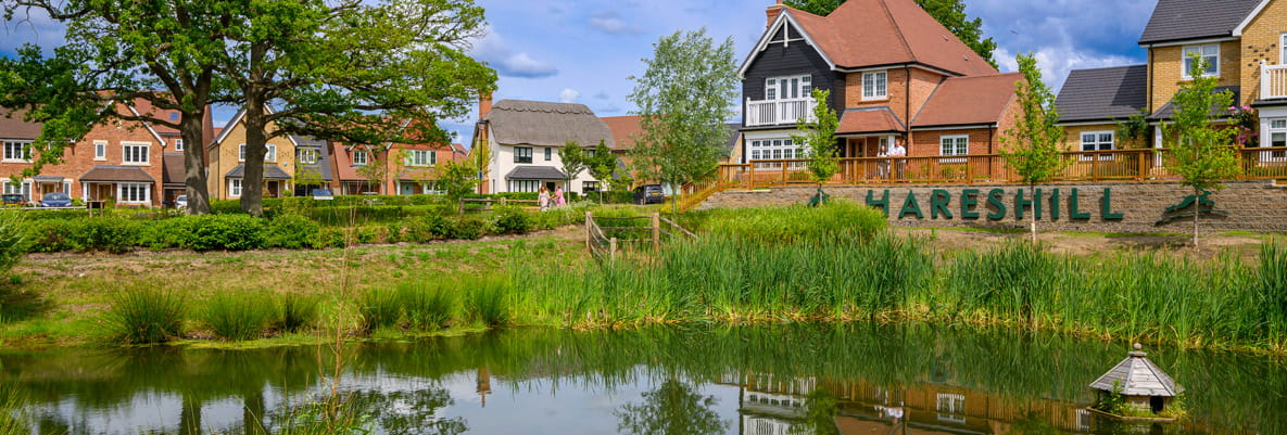Grass around clear pond with reflection of the new build development, a sign that reads Hareshill can be seen in background