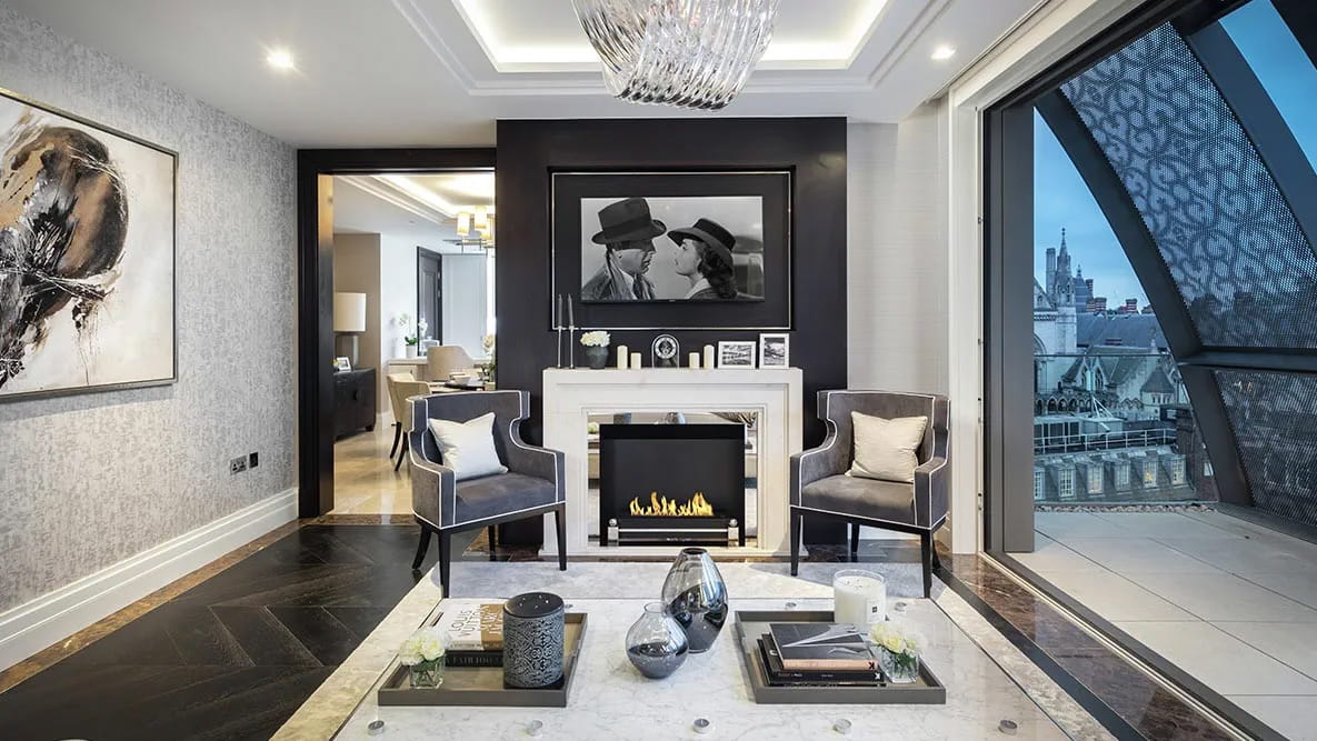 A wall-mounted television in a modern apartment. The television hangs above an electric fireplace with two grey armchairs in front.