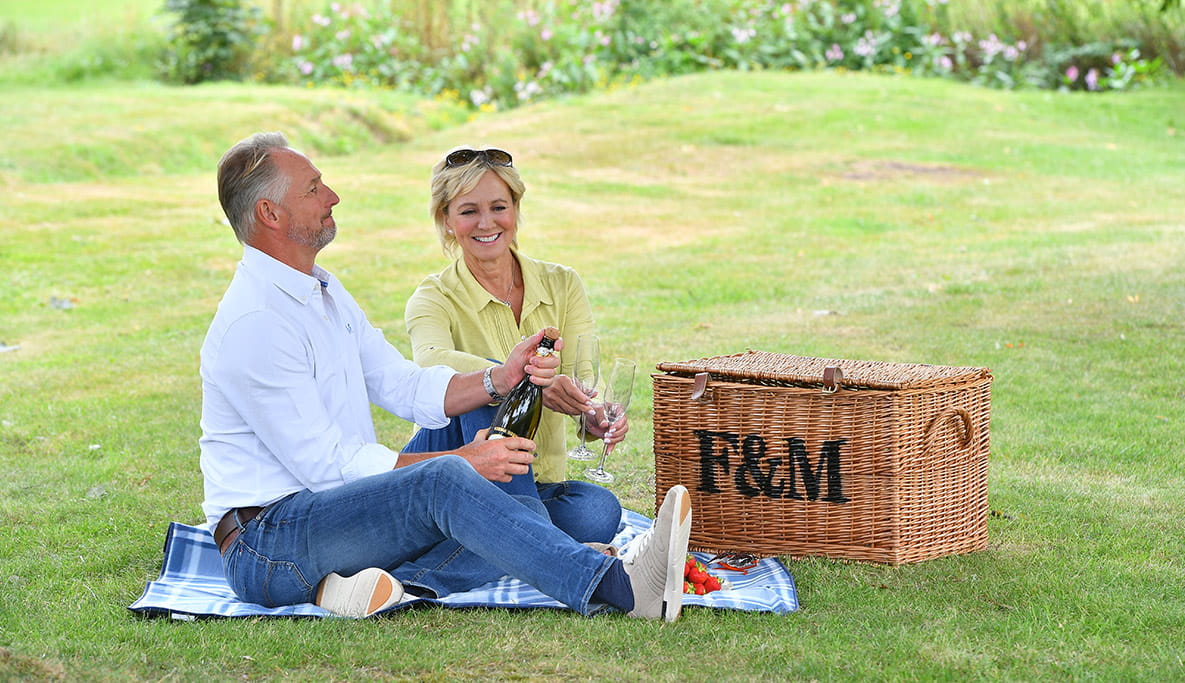 A couple sat having a picnic, opening a bottle of prosecco.