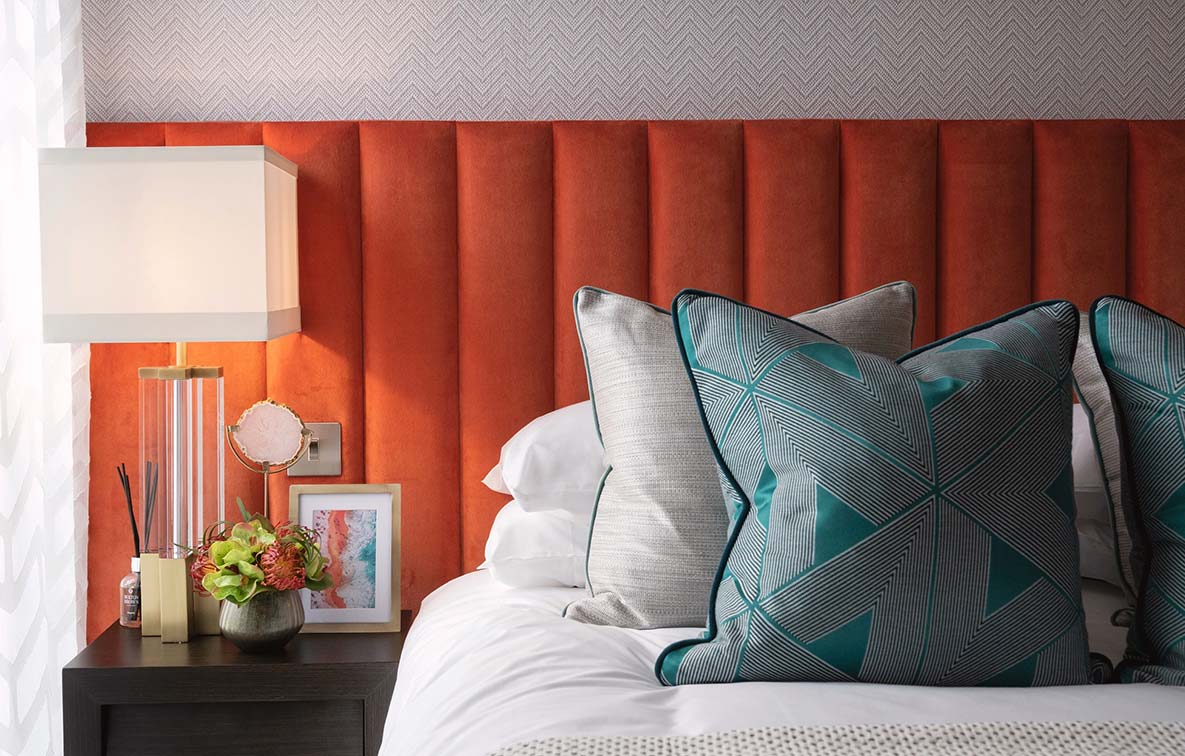 The corner of a bed, laden with colourful cushions to complement the bright orange headboard.