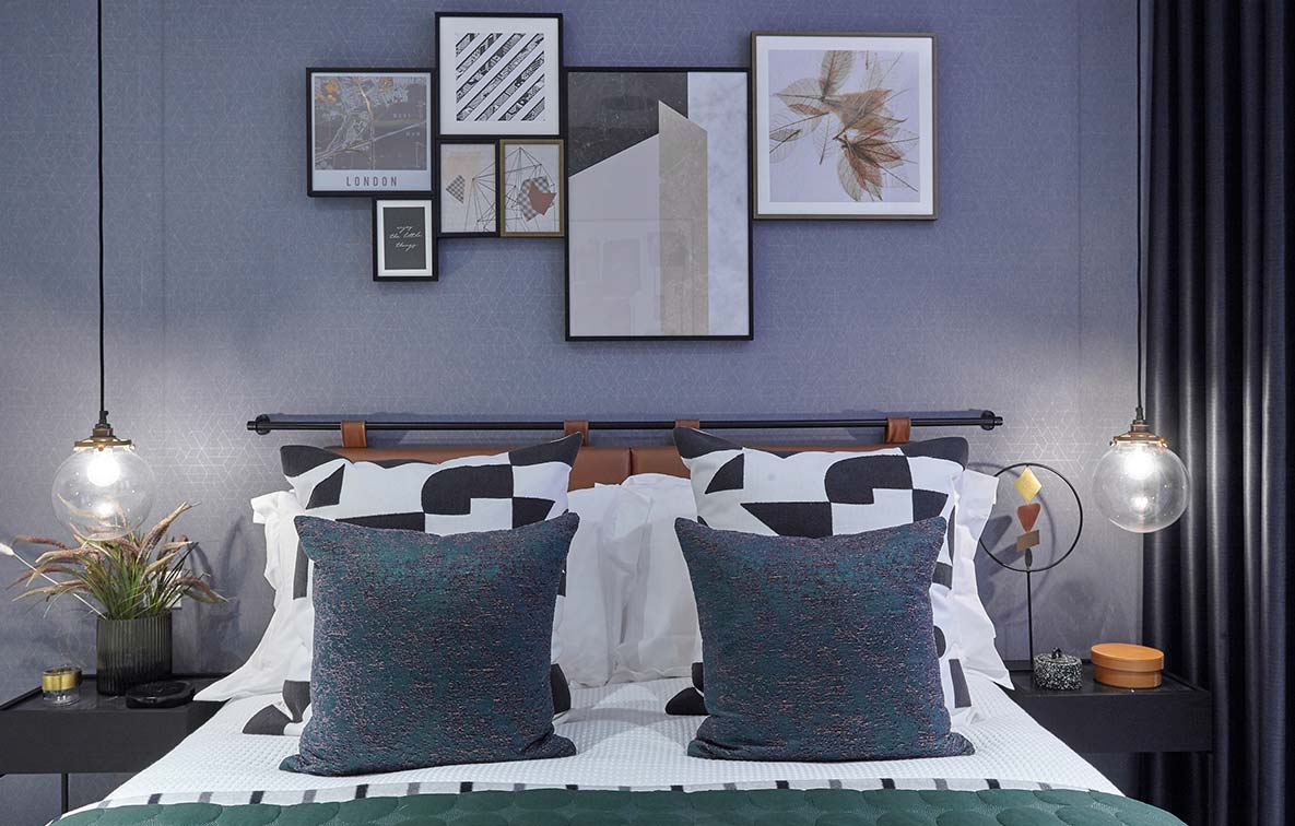 The head of a bed, complemented by a mixture of bright blue and geometric cushions, contemporary bedside tables and modern artwork. Low ceiling lights hang either side of the bed to create ambience in the room.