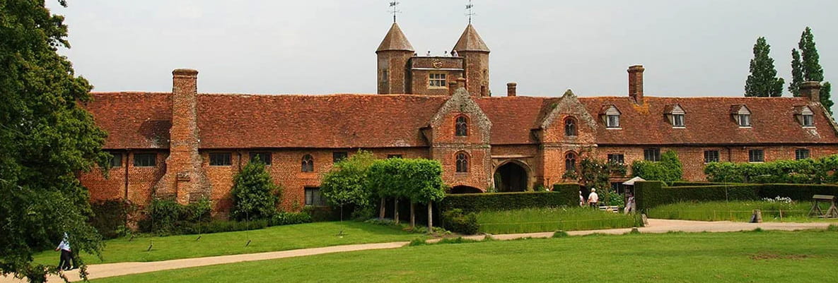 A historic brick building with a red-tiled roof. In front of the building is a green lawn with a few people walking around and trees in the background. 