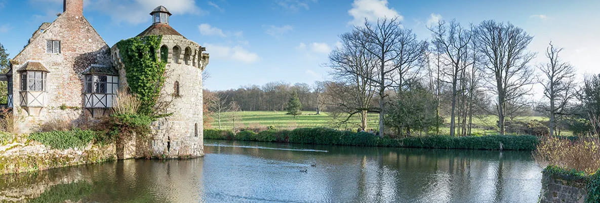 A castle with a moat. The castle is reflected perfectly in the surrounding water. In the background, the sky is blue and there’s a large field lined with trees. 