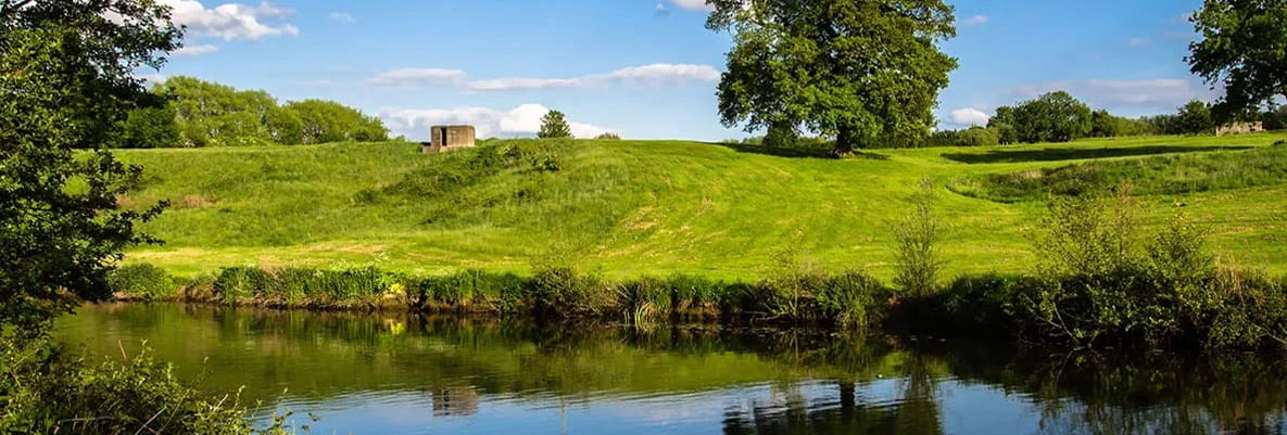 A river flowing through the green countryside on a sunny day. The river is clear and calm, with trees that line the river banks. 