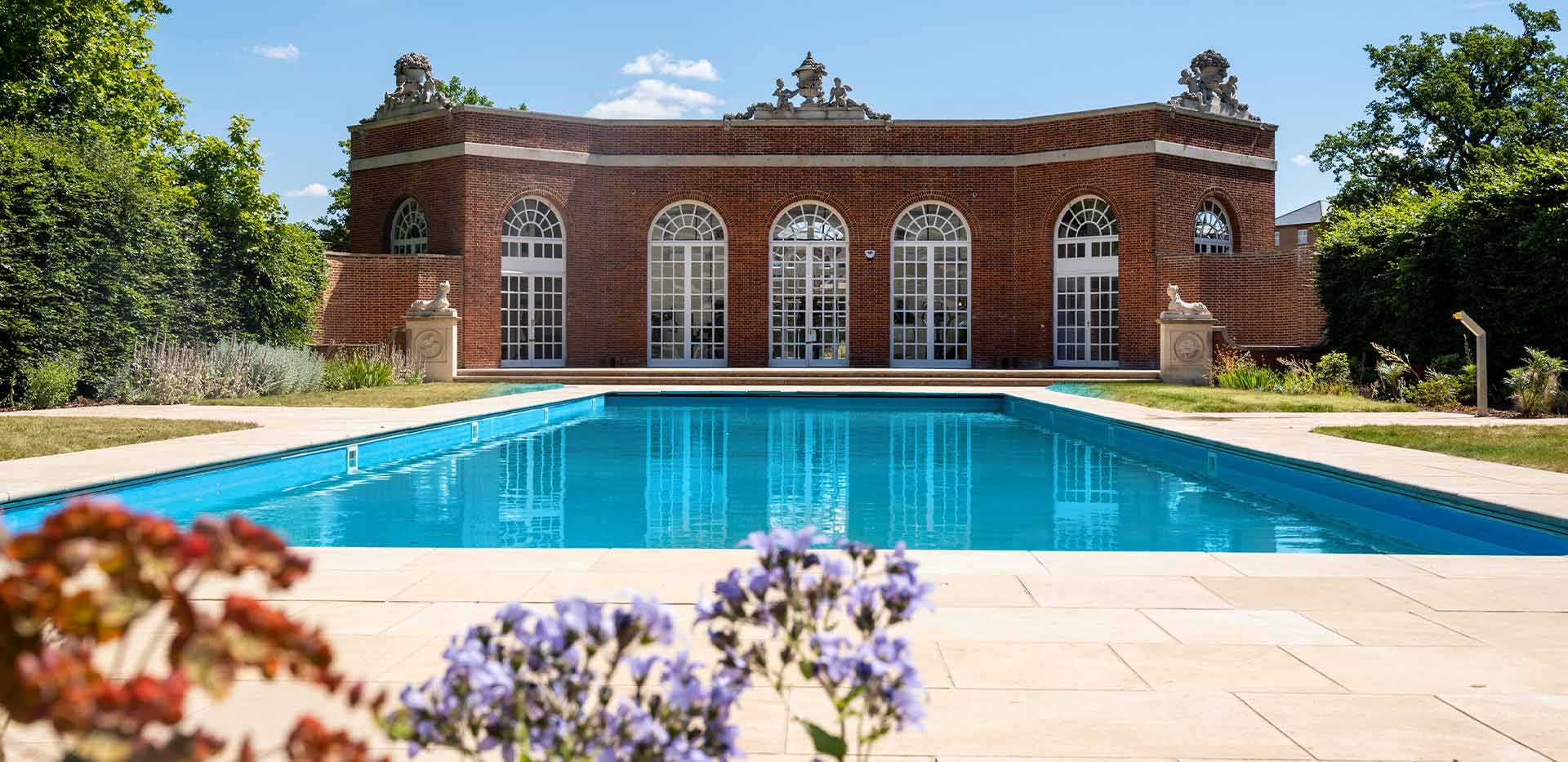 The 1920s-restored swimming pool at Trent Park, surrounded by pathways and flower beds, and overlooked by a red-brick building.