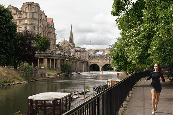 Female jogging along river or canalside against a backdrop of Bath city's buildings.