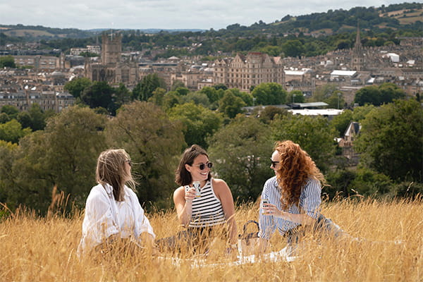 Three females sitting in tall yellow grass, against a background of Bath city and green hillsides.
