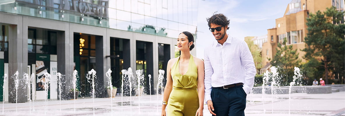 Female and male in smart attire, casually walking through a courtyard with fountains and building in background