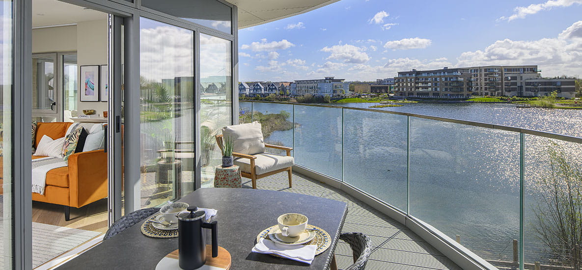 An apartment at Bankside Gardens. A large balcony overlooks a lake, with comfortable outdoor seating areas.