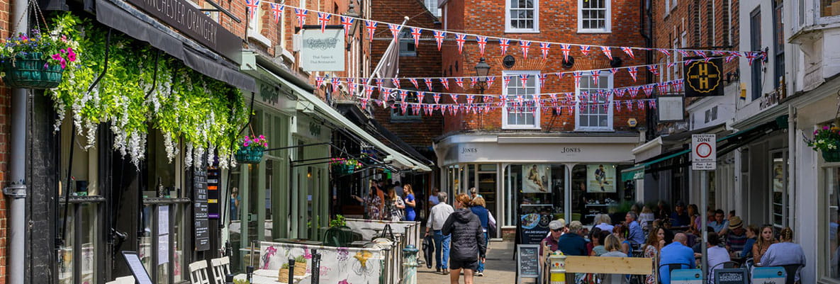 A street bustling with people eating, drinking and walking in Winchester. A selection of cafes and restaurants along the street provide outdoor seating areas for visitors to enjoy the historic setting.