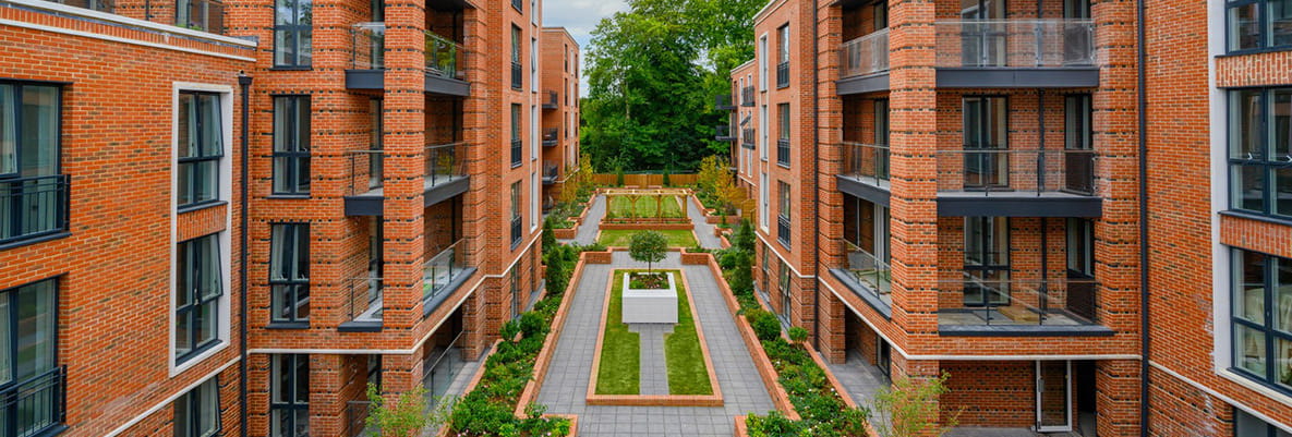 The exterior of a selection of apartment buildings at Knights Quarter—a development by Berkeley Group. The apartment buildings feature red brick and black accents around the windows and balconies. Along the paths leading to the buildings, large flowerbeds line the walkways. 