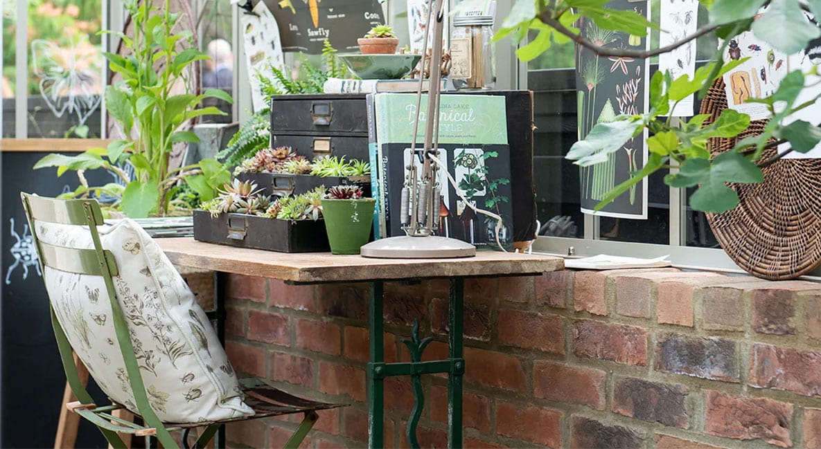 A small desk with plants, books and a lamp in a greenhouse.
