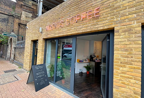 The exterior of Nostos, a coffee shop in Battersea. Featuring contemporary brickwork and black accent door frames, complete with blackboard advertising specials outside. 