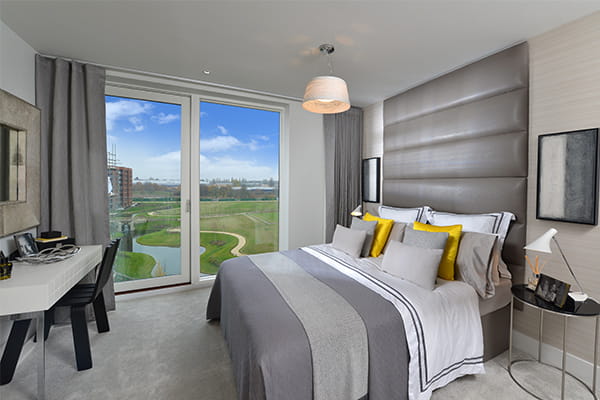 A stylish modern bedroom with a large bed adorned with yellow and grey cushions, a desk, and large windows overlooking a park.