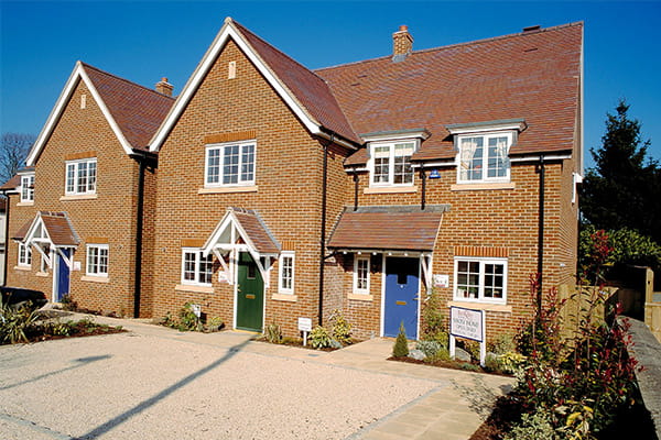 Two red brick semi-detached houses with pitched roofs and blue and green front doors, surrounded by gardens and a gravel driveway.