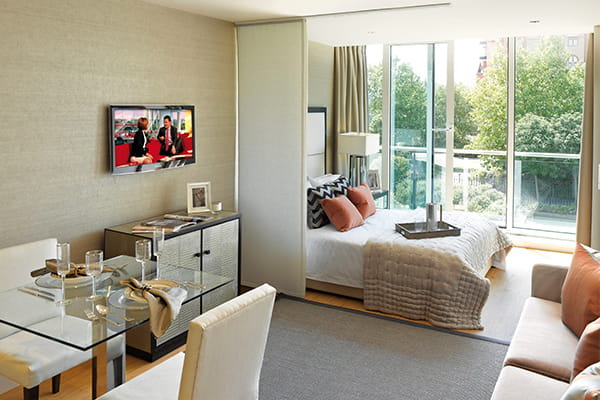 A modern studio apartment with a dining area, a sofa, a bed, and large windows offering a view of trees outside.