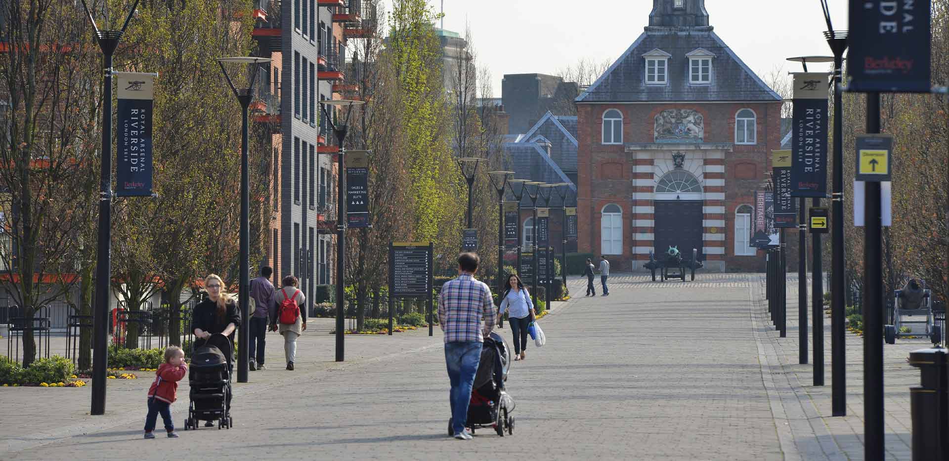 Residents and visitors wandering around Royal Arsenal Riverside—a development by Berkeley Group. Lamposts with banners line the large pavements, while pedestrians go about their daily business.