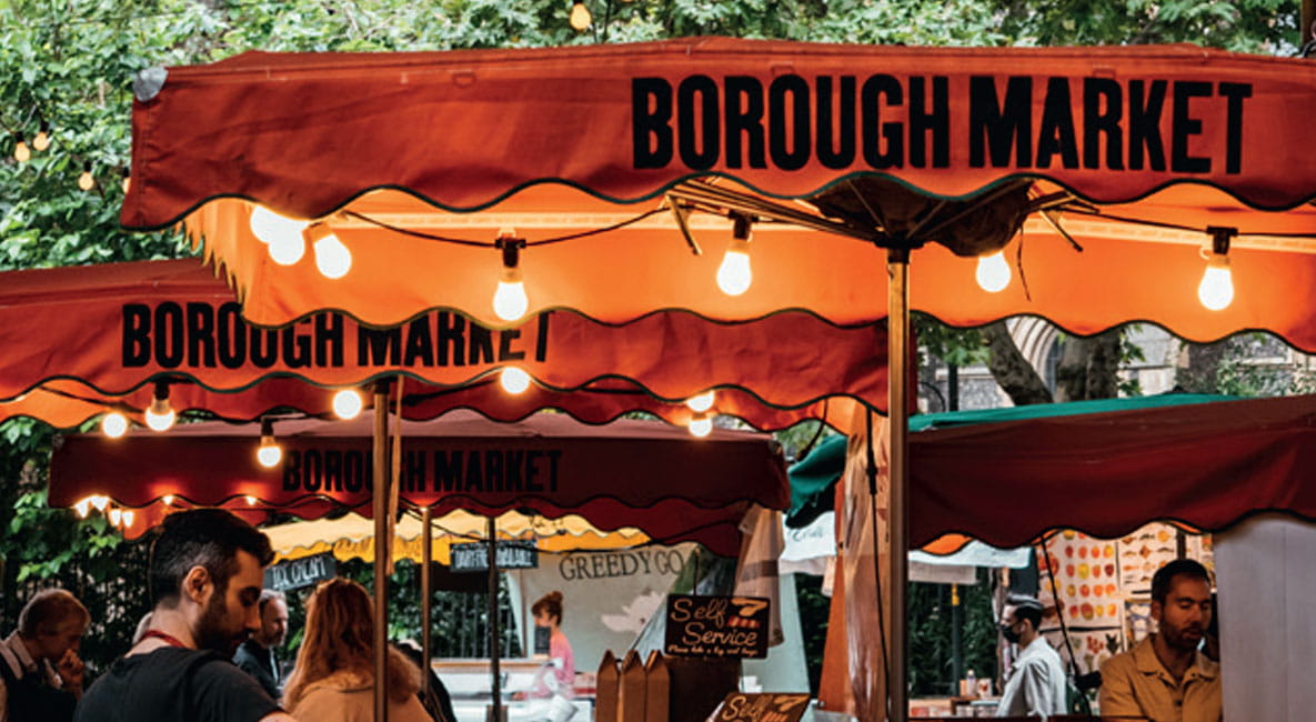 Borough market. Several people are walking around looking at the stalls which sit under orange canopies with ‘Borough Market’ written on them.
