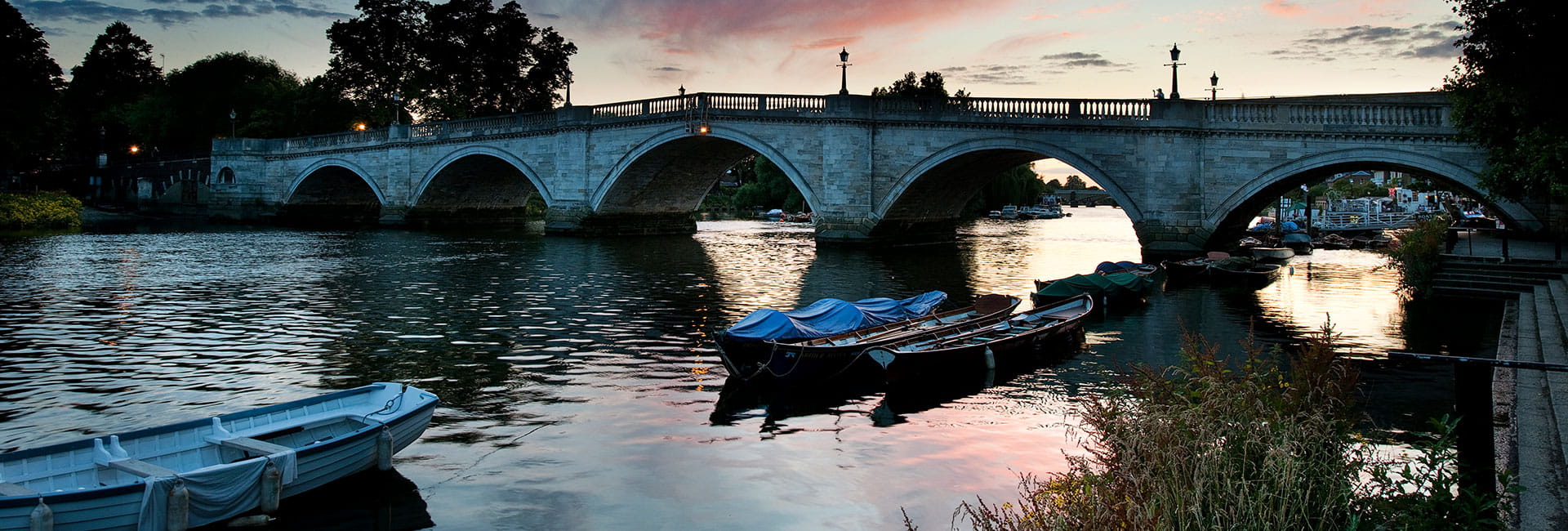 A scenic river view in Surrey, featuring boats on the water and a large bridge in the background, providing access to either side of the river. In the distance, the sun sets behind the bridge and reflects on the water.
