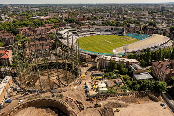 Aerial view of a construction site near a large stadium and surrounding urban landscape under a blue sky with scattered clouds.