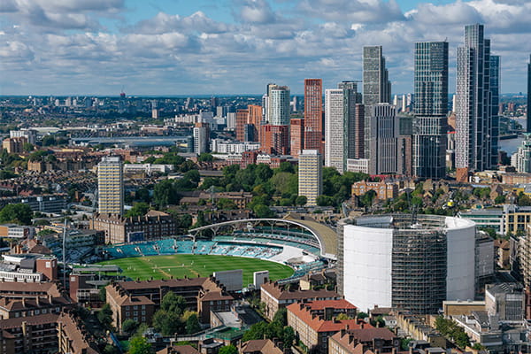 A cricket ground with green seating, a scoreboard, and a backdrop of buildings and a historic gasholder structure on a sunny day.