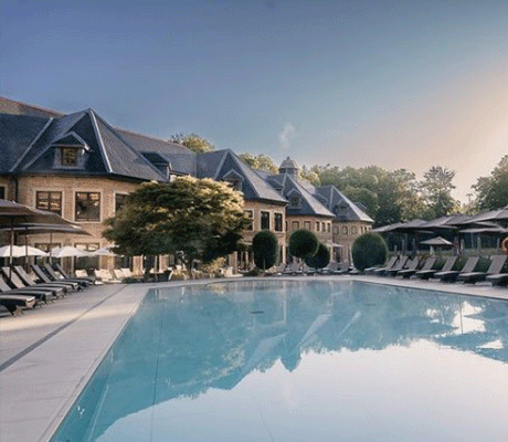 An outdoor swimming pool lined with sun loungers. The building of Pennyhill Park luxury spa hotel overlooks the outdoor pool.