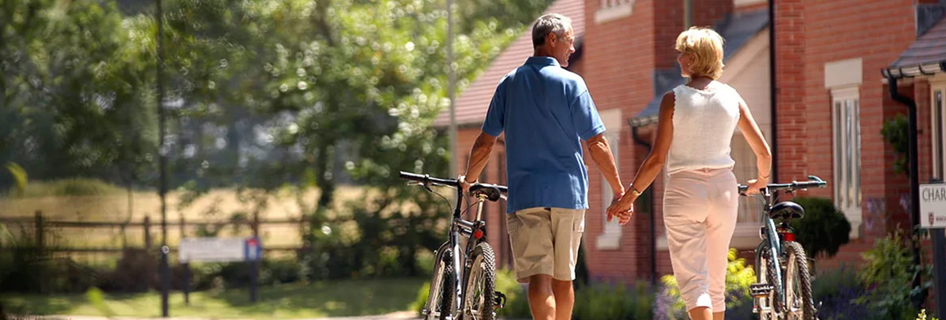 A couple hold hands as they walk along a public footpath, surrounded by residential housing and green space. They each hold a bicycle with their other hand, which they wheel alongside them.