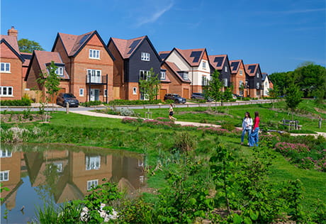 people walking along a path beside green landscaped areas and water feature with new build homes in the background