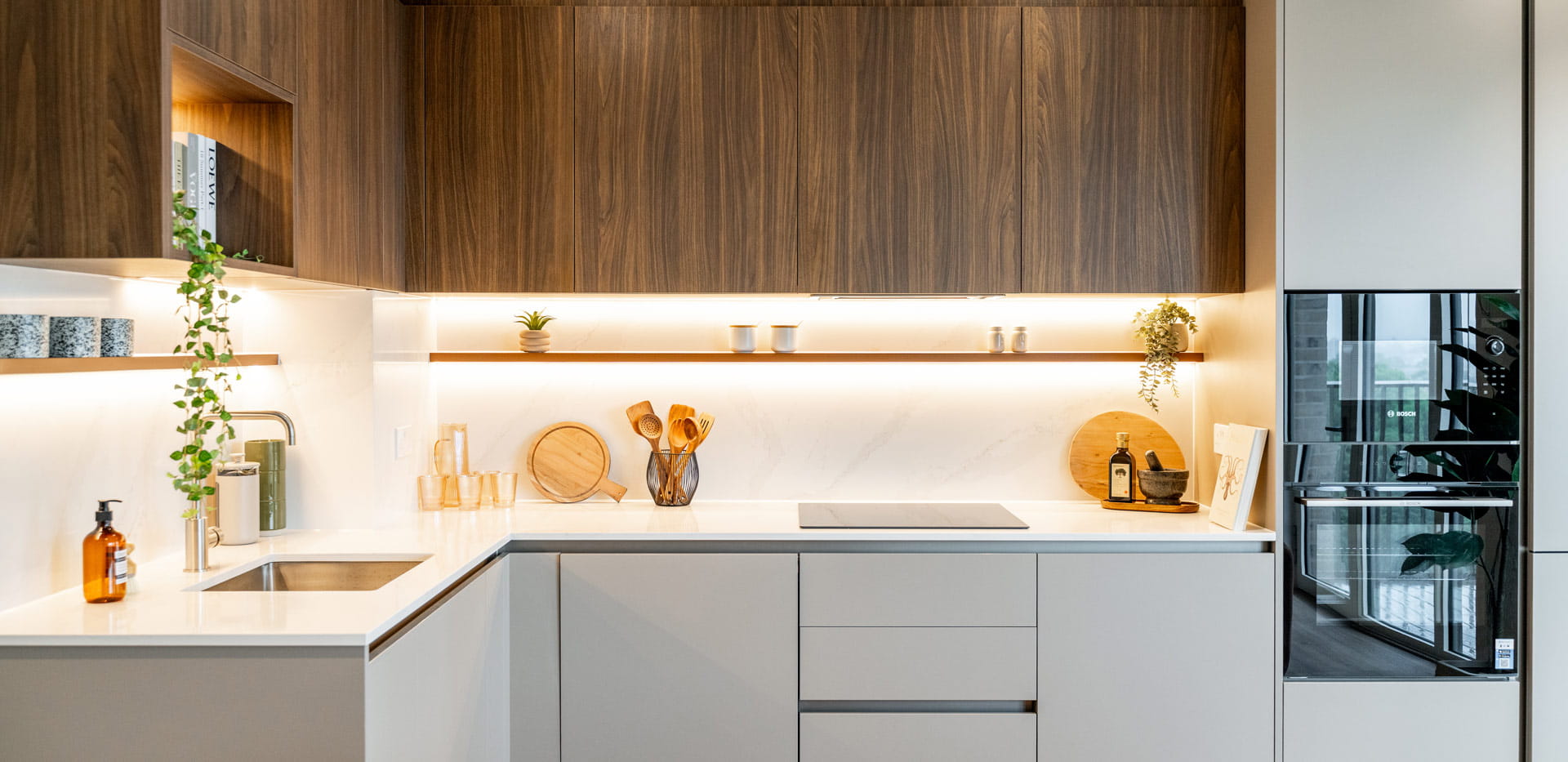 View of kitchen with modern appliances, white counters, chrome taps, strip lighting, overhead cupboards.