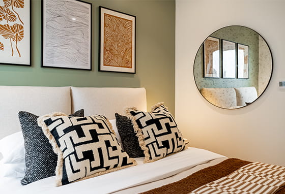 Closeup view of a made up bed, with wall hangings above and circular wall hanging mirror.