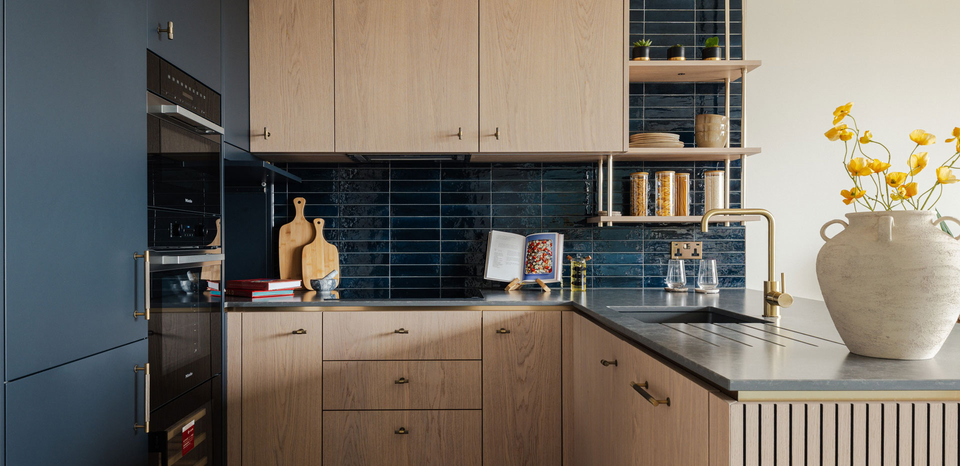 Kitchen with attached island, dark blue splashback tiles, light wood brown cupboards, grey countertops.