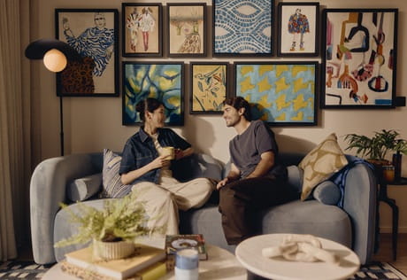 A young couple sitting on fabric sofa, abstract wall hangings above, around modern decorated living space.