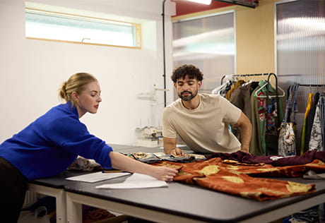 Two people examining clothing fabric sprawled on a wide table.