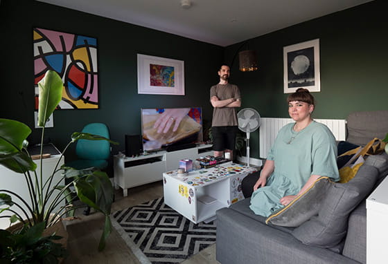 A couple of posing for a photo in their living room. A woman is sitting on her sofa with her partner standing up in the background.
