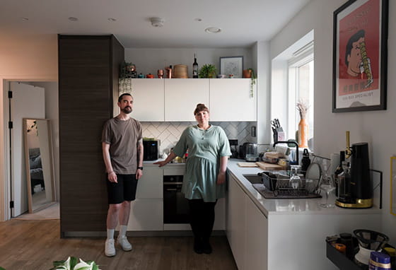 A couple are in their kitchen posing for a photo. Their kitchen is spacious with a large window, causing natural light to filter in.