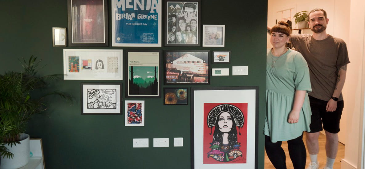 A couple are posing for a photo in their hallway, next to an impressive canvas collection displayed on their living room wall.