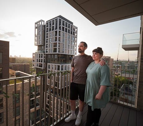 A couple are admiring the view of Kidbrooke Village from their high-rise apartment balcony. The sun is setting in the background. 