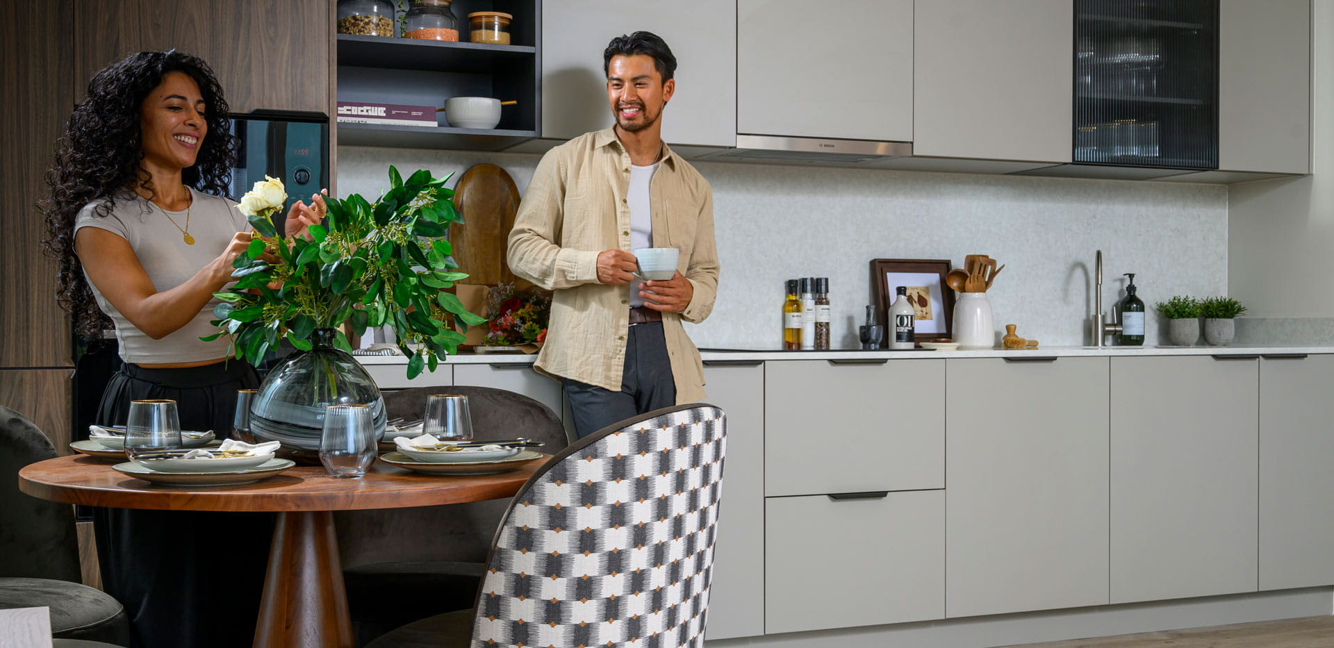 Male holding cup smiling and facing female decorating flower arrangement in dining space.