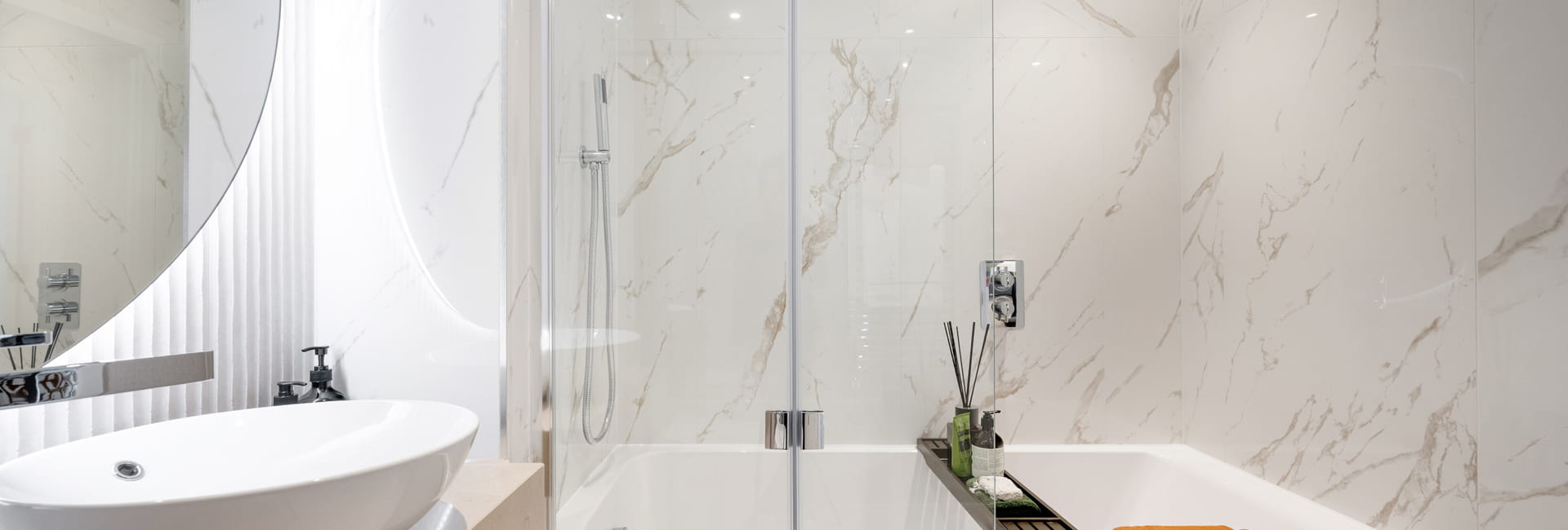 Shower over bath, wash basin with mirror and vanity lighting, marble texture white tiles.