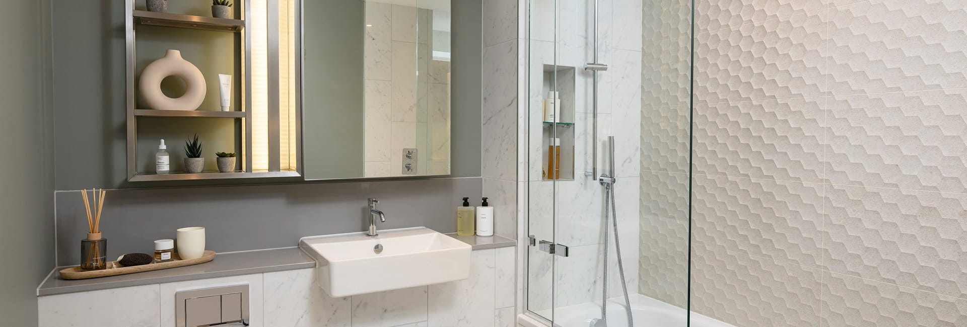 Bathroom with shower over bath, beige honeycomb style tiling, wash basin with mirror, shelving.