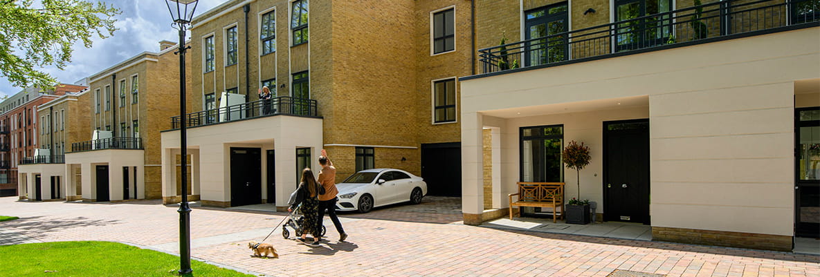 Couple with pram and dog walking along quiet road pavement beside new build homes waving to a neighbour on balcony