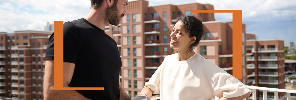 Male and female enjoying a beverage and conversation out on a balcony, on a sunny day with apartment blocks in background.