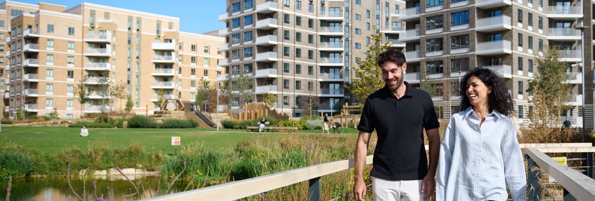 Two people walking on a footbridge beside a pond, with modern flats and green space behind them.