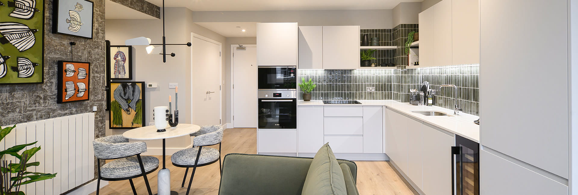 Modern open-plan flat with white kitchen, round dining table, green sofa and framed artwork on the wall.