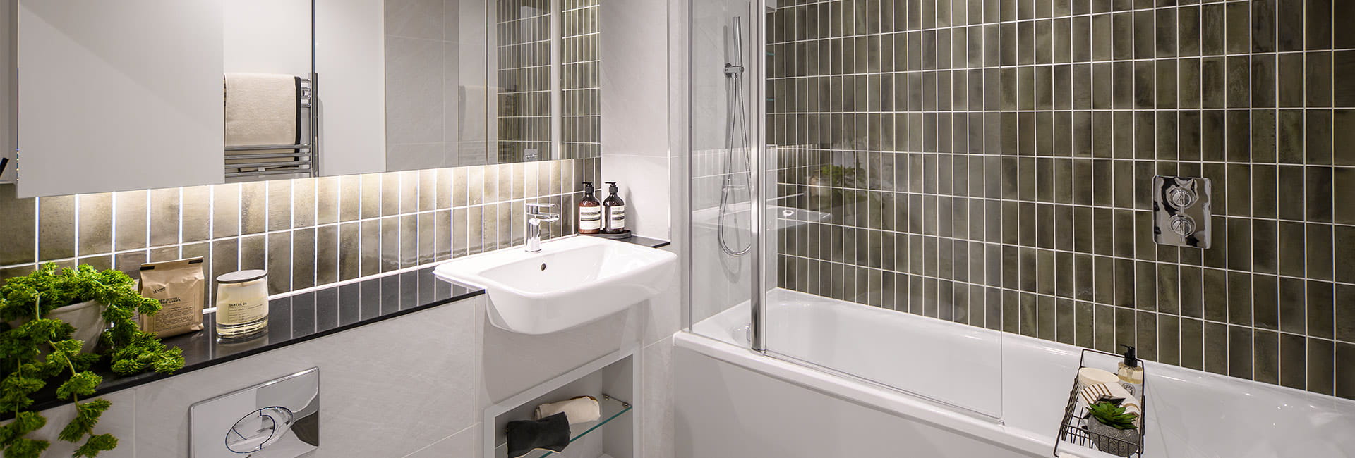 Modern bathroom with mirrored cabinets, small sink, bath with shower screen, and dark olive tiled wall.