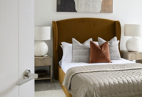 Closeup of bedside with tan-brown fabric velvet headboard, wall art against off-white wall, designer lamp and bedside table.
