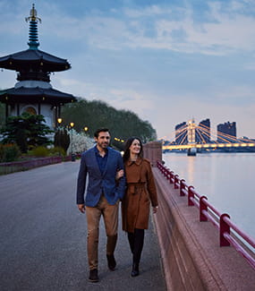 A couple walking in Battersea Park at dusk with Chelsea Bridge in the background